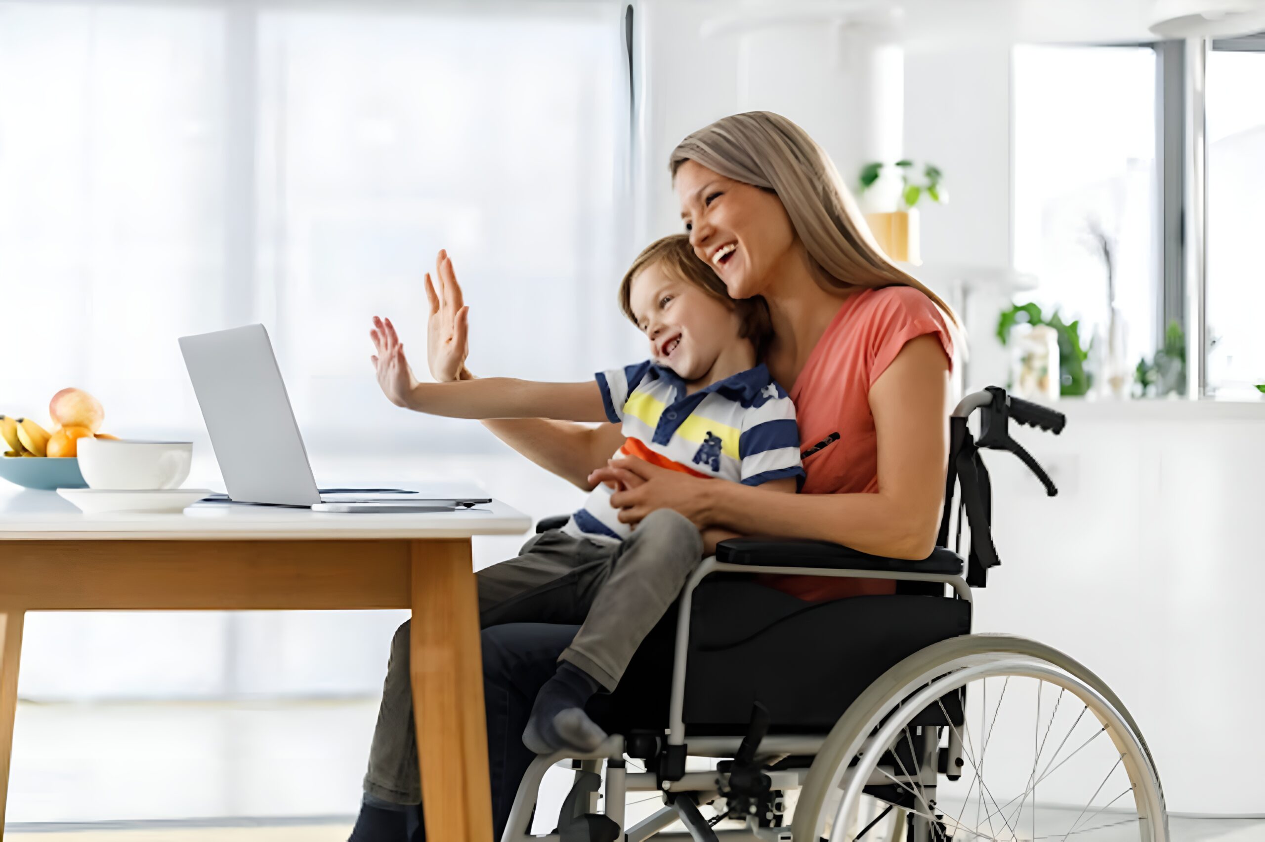 A person in a wheelchair with a toddler in their lap. They are waving at a laptop in front of them which suggests they might be on a video call.