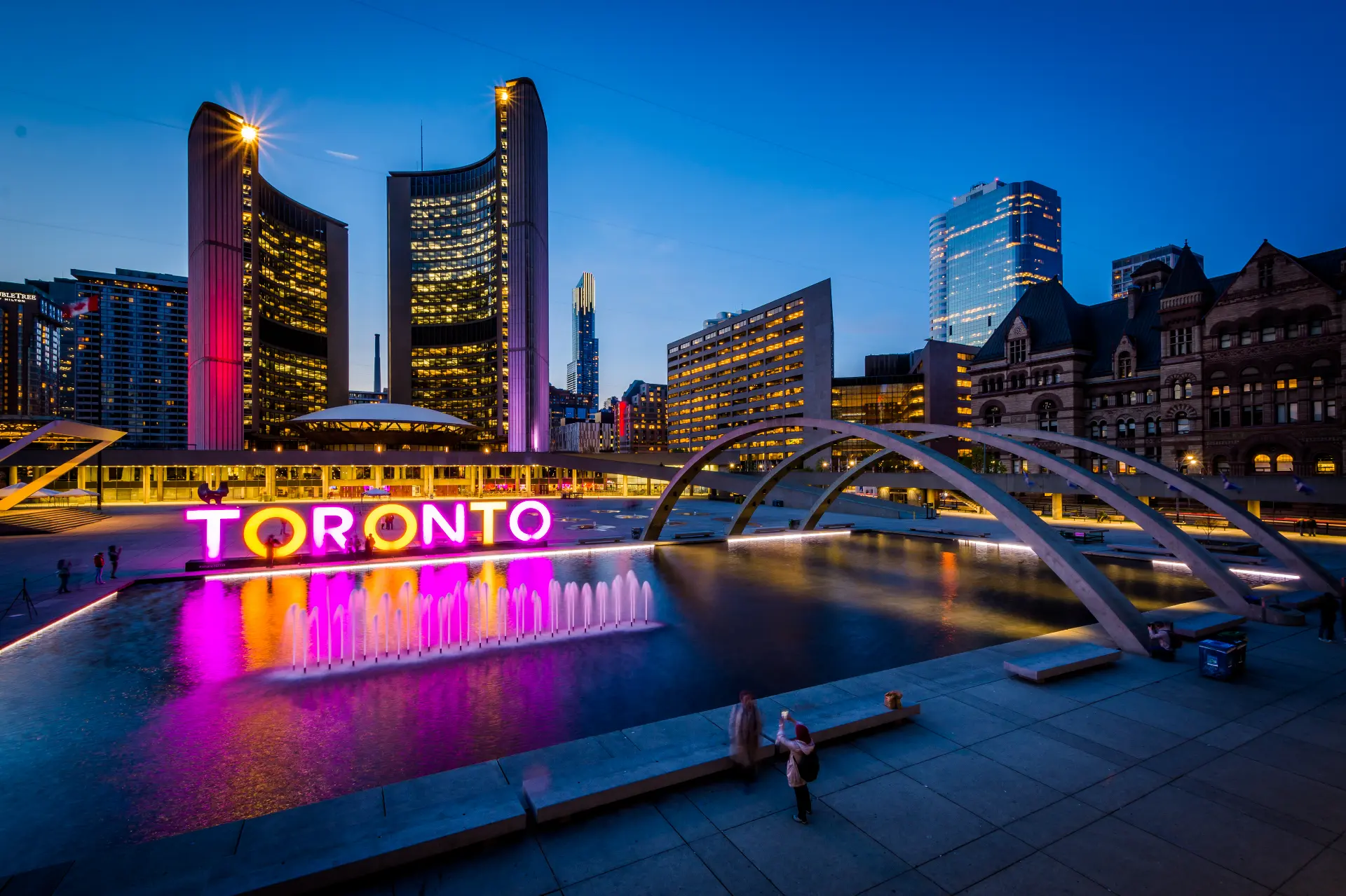 An evening picture of Toronto city hall with both the lighted Toronto logo and an artificial pond in the foreground, in the who we are section.